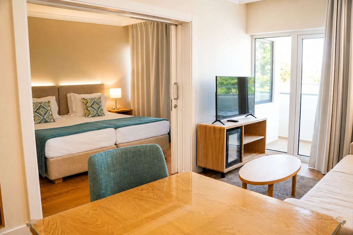 A hotel room with a bed, a TV, a dining table, and a view of the outdoors.