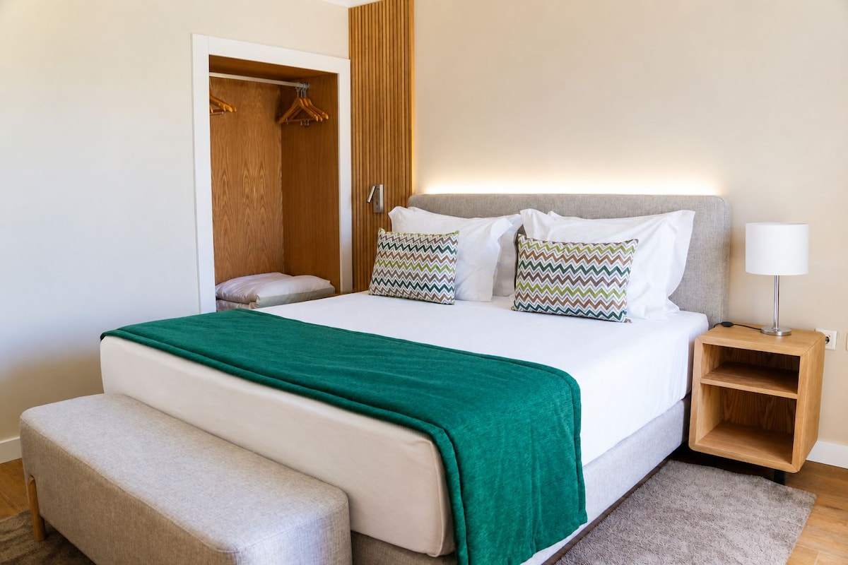 A neatly made bed with a green throw, a bedside table with a lamp, and a wooden wardrobe.