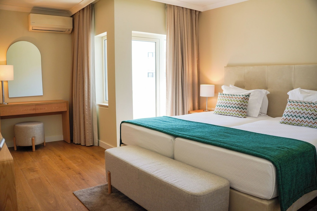 A hotel room with a bed, bedside lamps, a mirror, a small stool, and a window with curtains.
