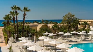 A resort pool area with white umbrellas and lounge chairs, palm trees, and a view of the sea.
