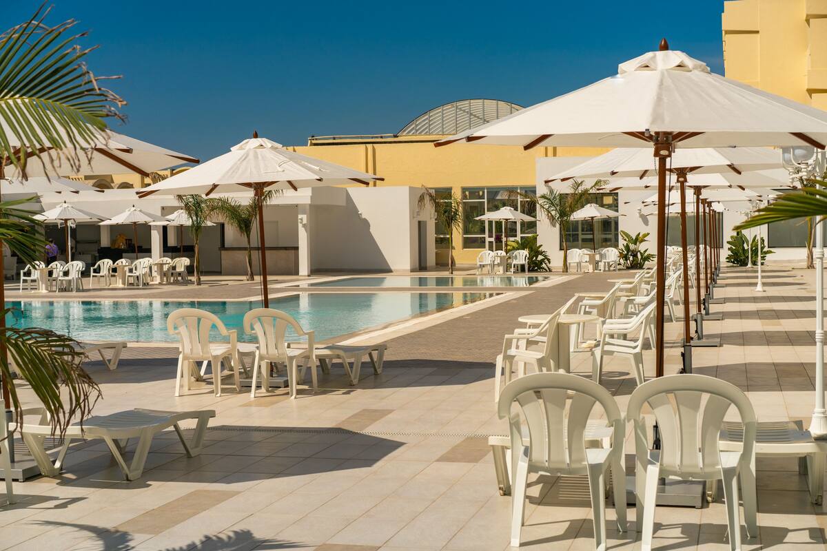 Zona de piscina con sillas y sombrillas blancas, cielo azul despejado y un edificio con fachada amarilla.