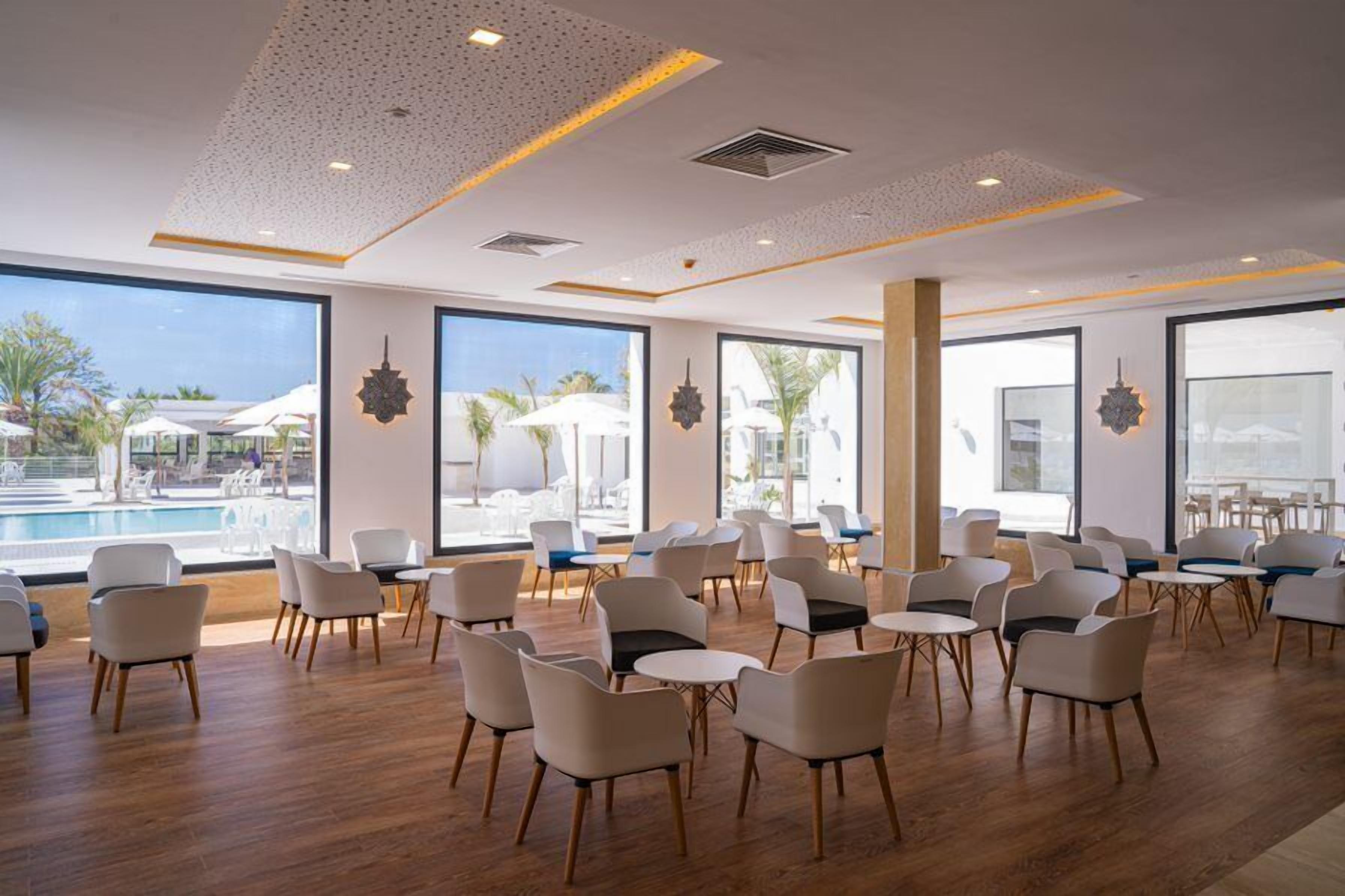 A spacious indoor area with multiple seating arrangements, large windows offering views of an outdoor pool area, and decorative wall elements.