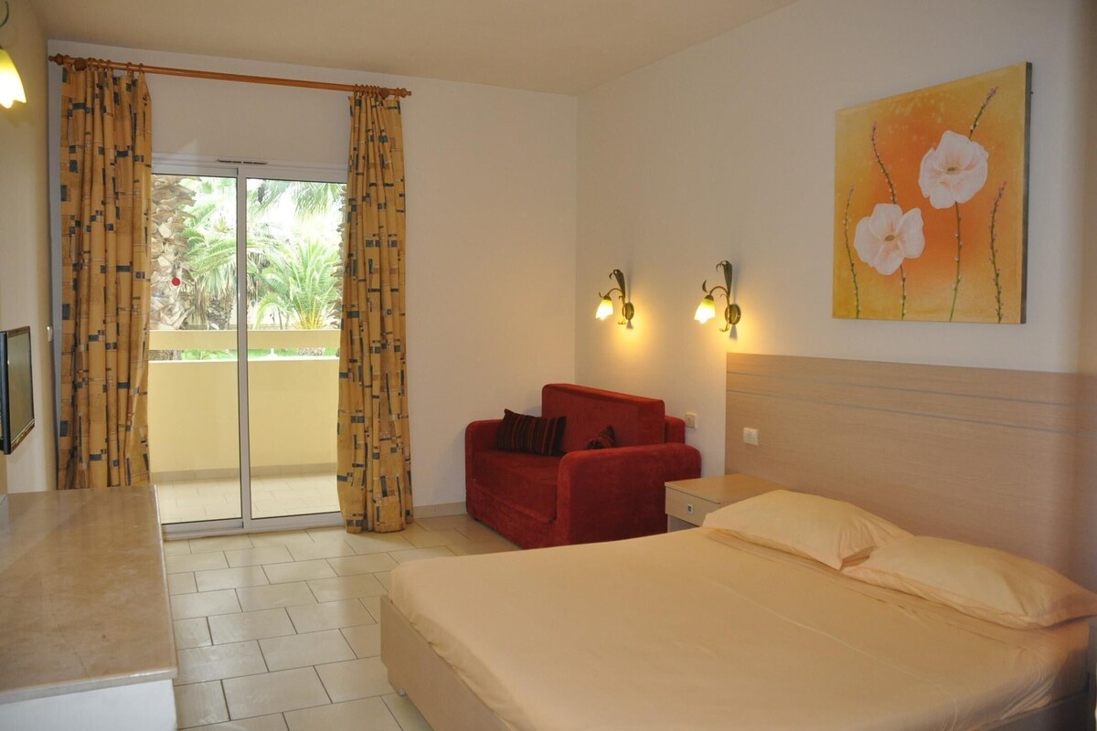 A bedroom with a bed, a red armchair, a painting, and a sliding glass door to a balcony.