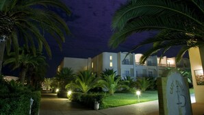 Vista nocturna de un hotel con palmeras y un camino iluminado que conduce a la entrada.