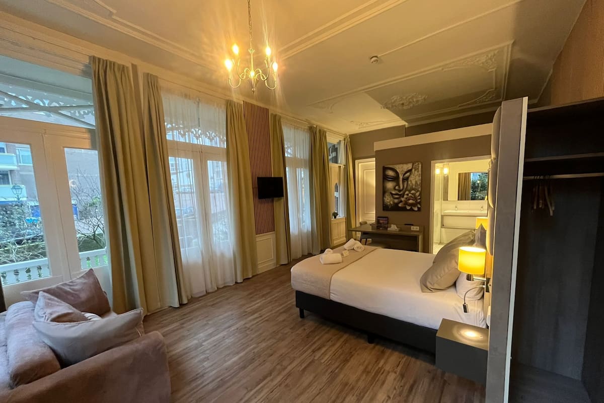 A hotel room with a bed, bedside table, lamp, TV, and a view of the outside.