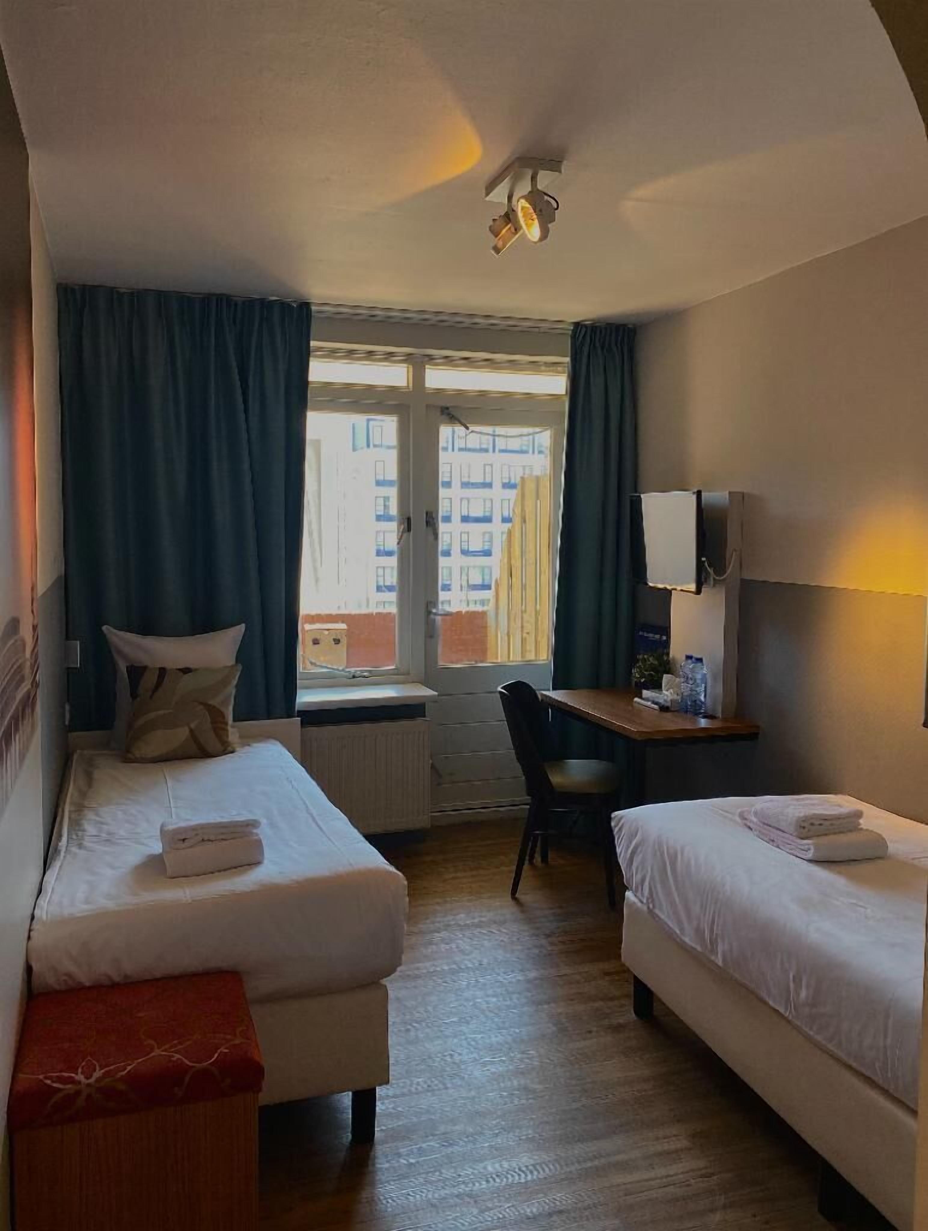 Une chambre d’hôtel avec deux lits, un bureau, une chaise et une fenêtre avec des rideaux.