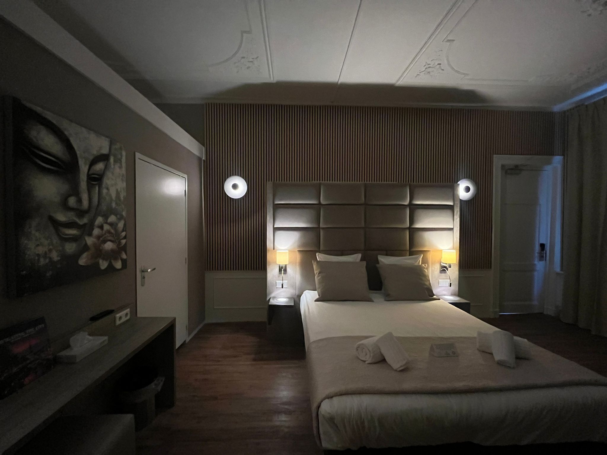 Habitación de hotel con una cama grande, dos lámparas de noche, cabecero con listones verticales, un cuadro en la pared y suelo de madera.