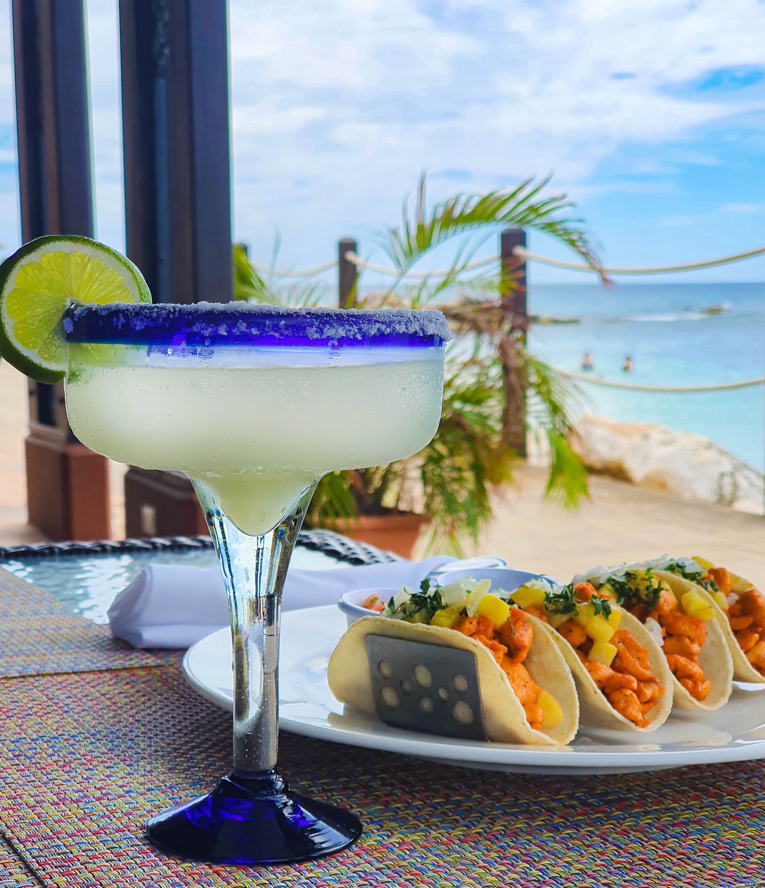 Lasissa on suolalla reunustettu reunus, ja siinä on limeviipaleella koristeltu margarita-cocktail. Edessä on lautanen täynnä tacoja, ja taustalla näkyy ranta.
