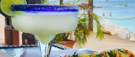 Un cóctel margarita con una rodaja de lima, un plato de tacos y una vista a la playa al fondo.