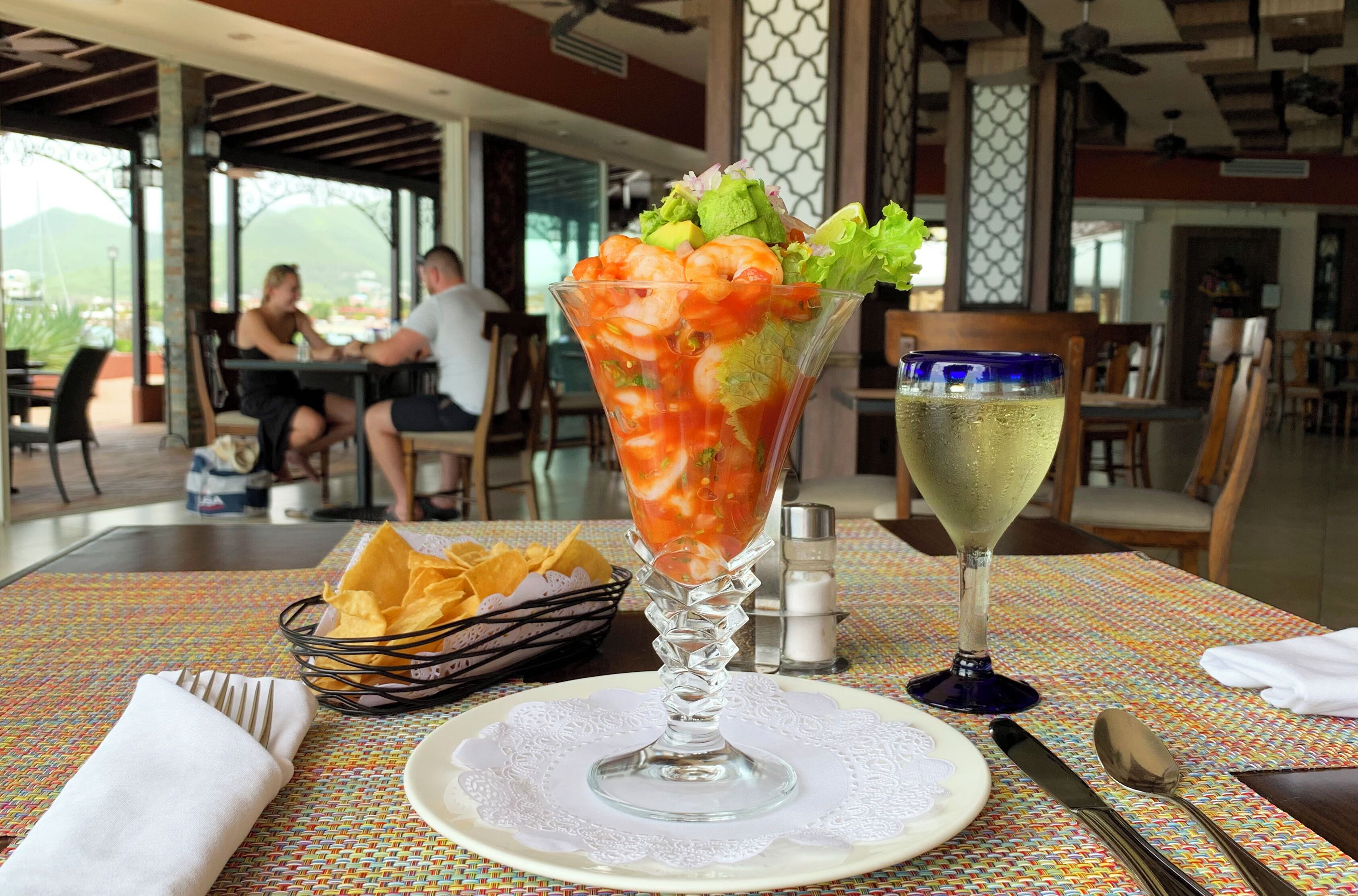 A dining table with a shrimp cocktail, a glass of white wine, and a basket of chips.