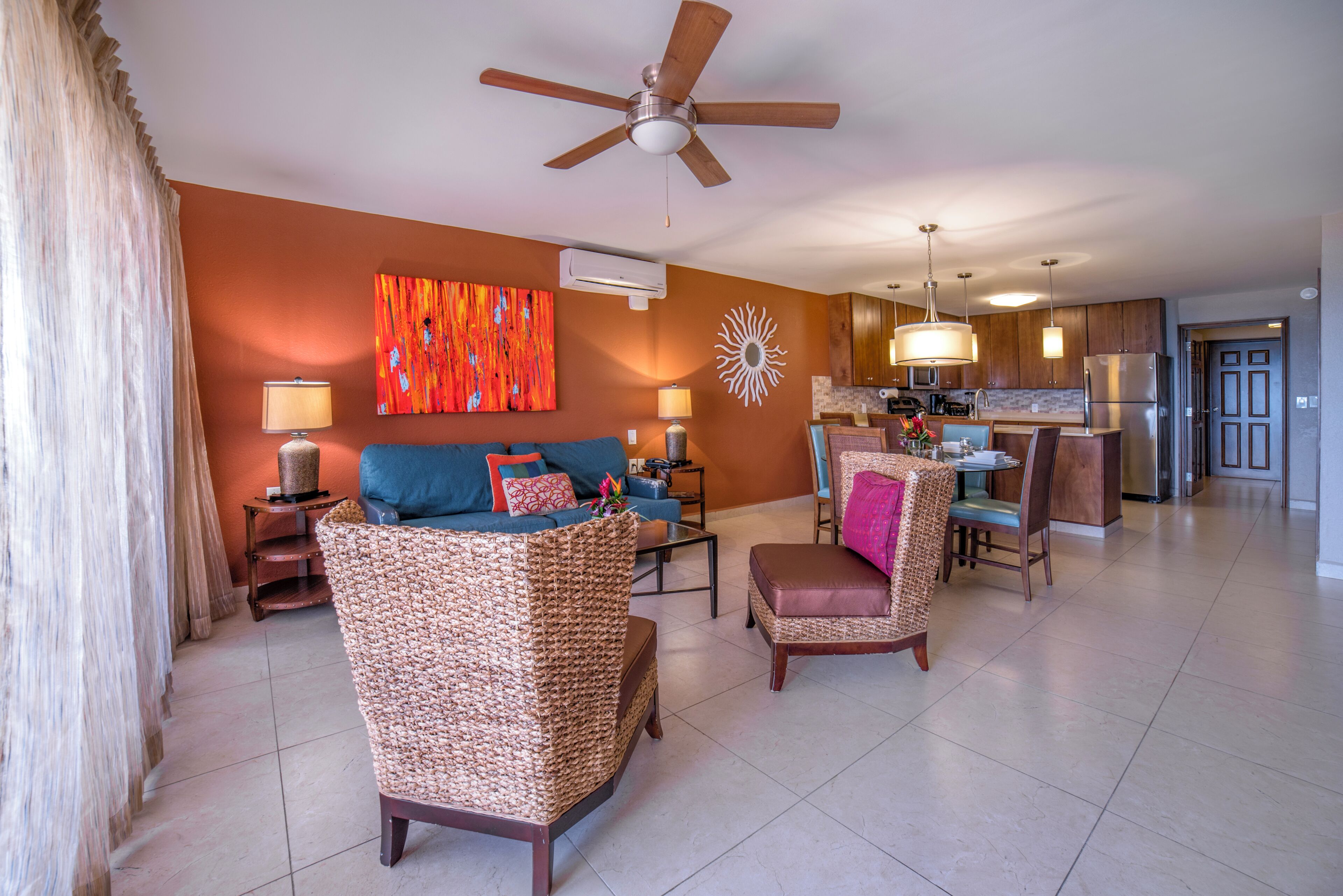 A spacious living area with a sofa, armchairs, and a dining table. A ceiling fan and an air conditioning unit are visible.