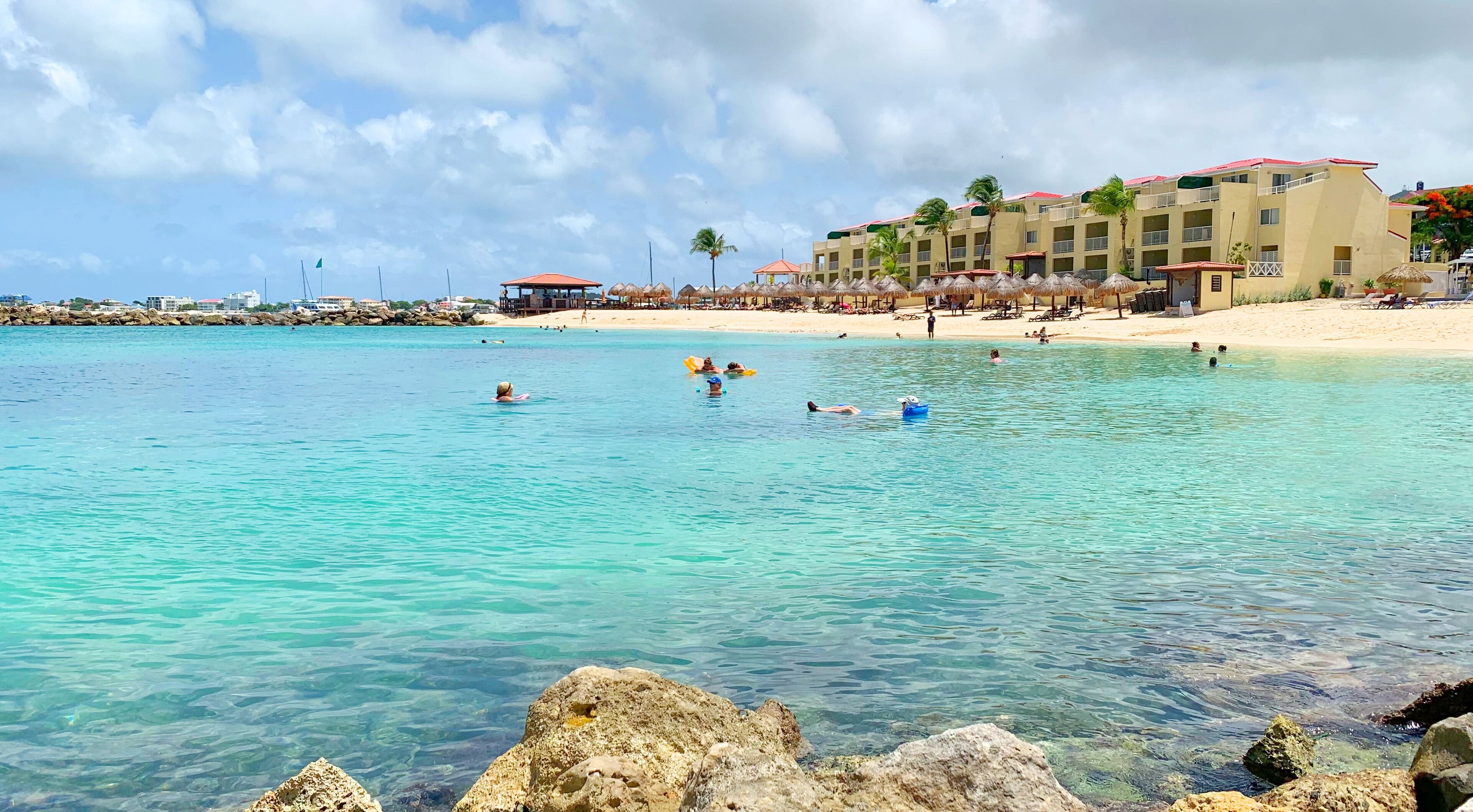 A beach resort with clear turquoise waters, a rocky shoreline, and a building with balconies overlooking the beach.