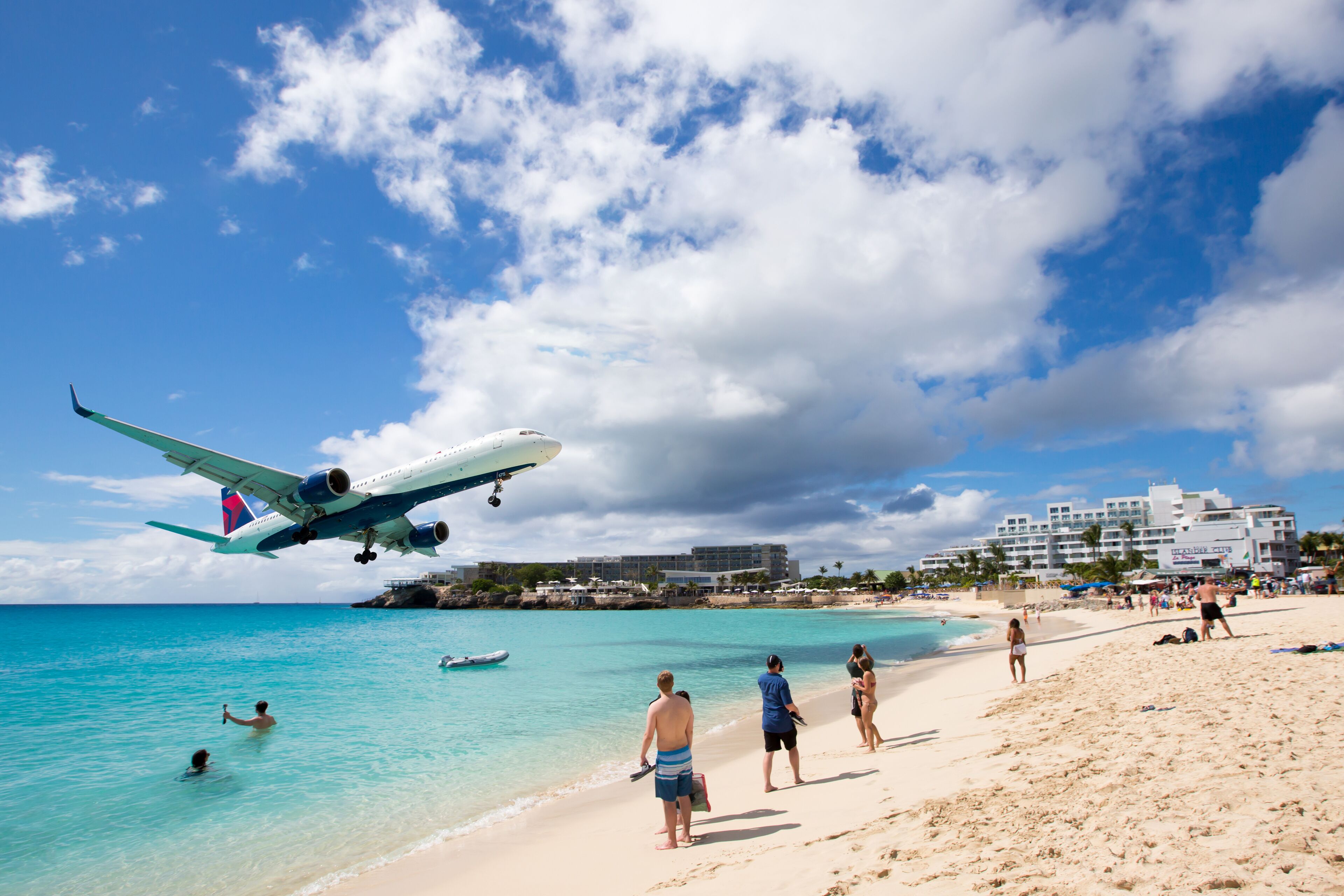 Una playa con gente, un avión aterrizando y un hotel al fondo.