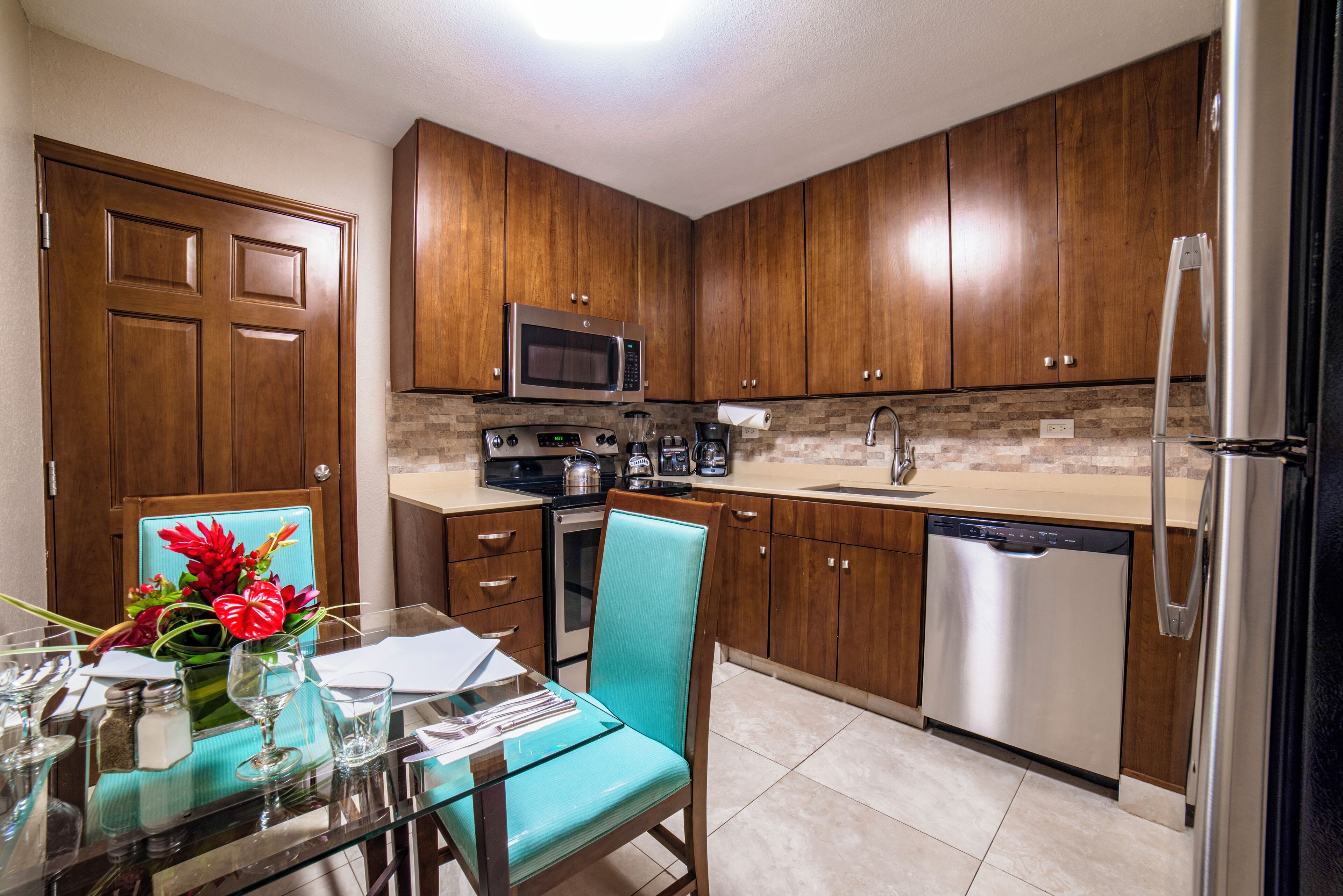 Una cocina moderna con armarios de madera, electrodomésticos de acero inoxidable y una zona de comedor con mesa de vidrio y una silla color turquesa.