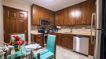 Una cocina moderna con armarios de madera, electrodomésticos de acero inoxidable y una zona de comedor con mesa de vidrio y una silla color turquesa.