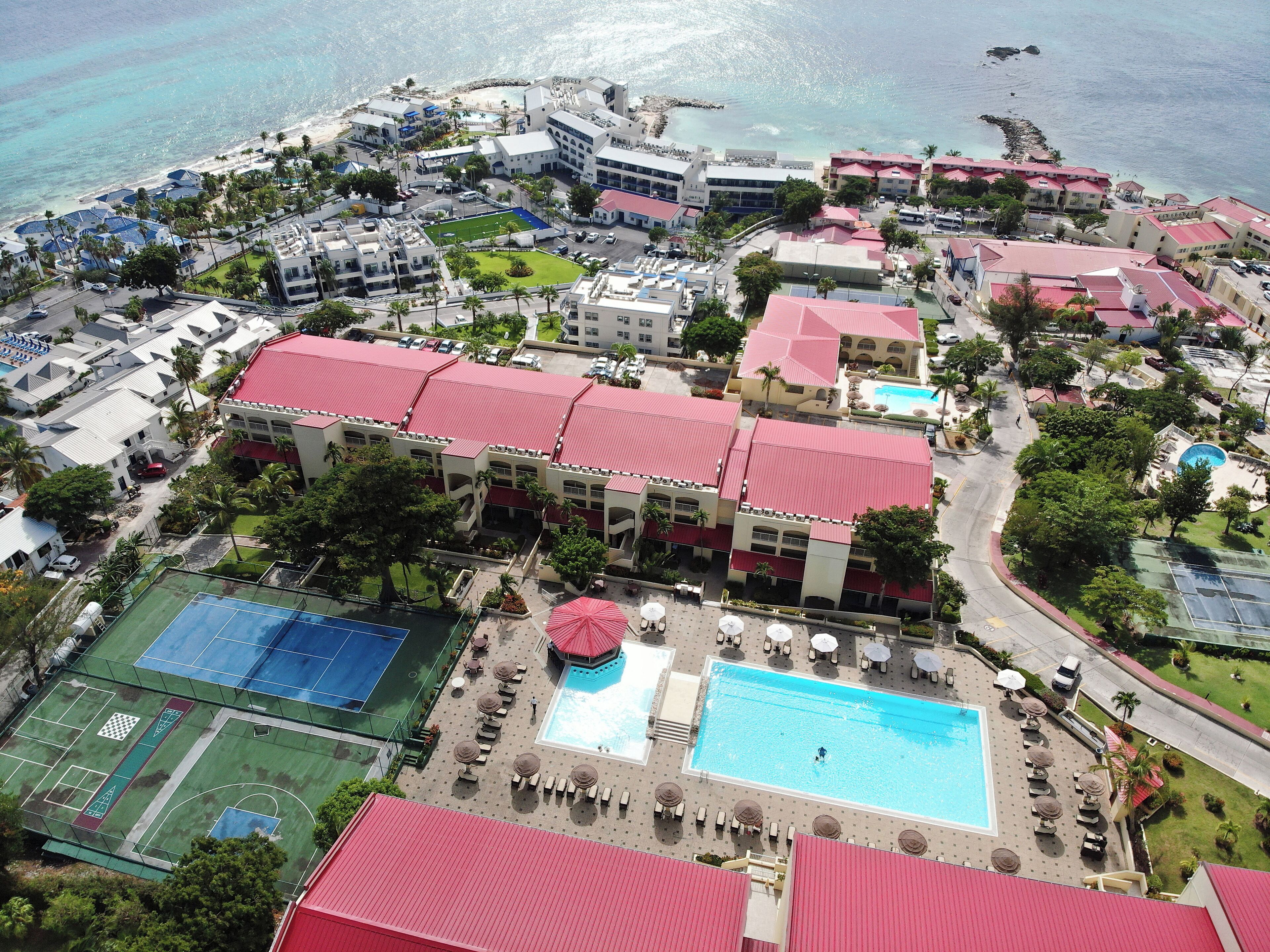 An aerial view of a resort with multiple buildings, a large swimming pool, tennis courts, and a beach area.