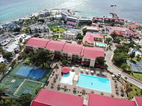 An aerial view of a resort with multiple buildings, a large swimming pool, tennis courts, and a beach area.