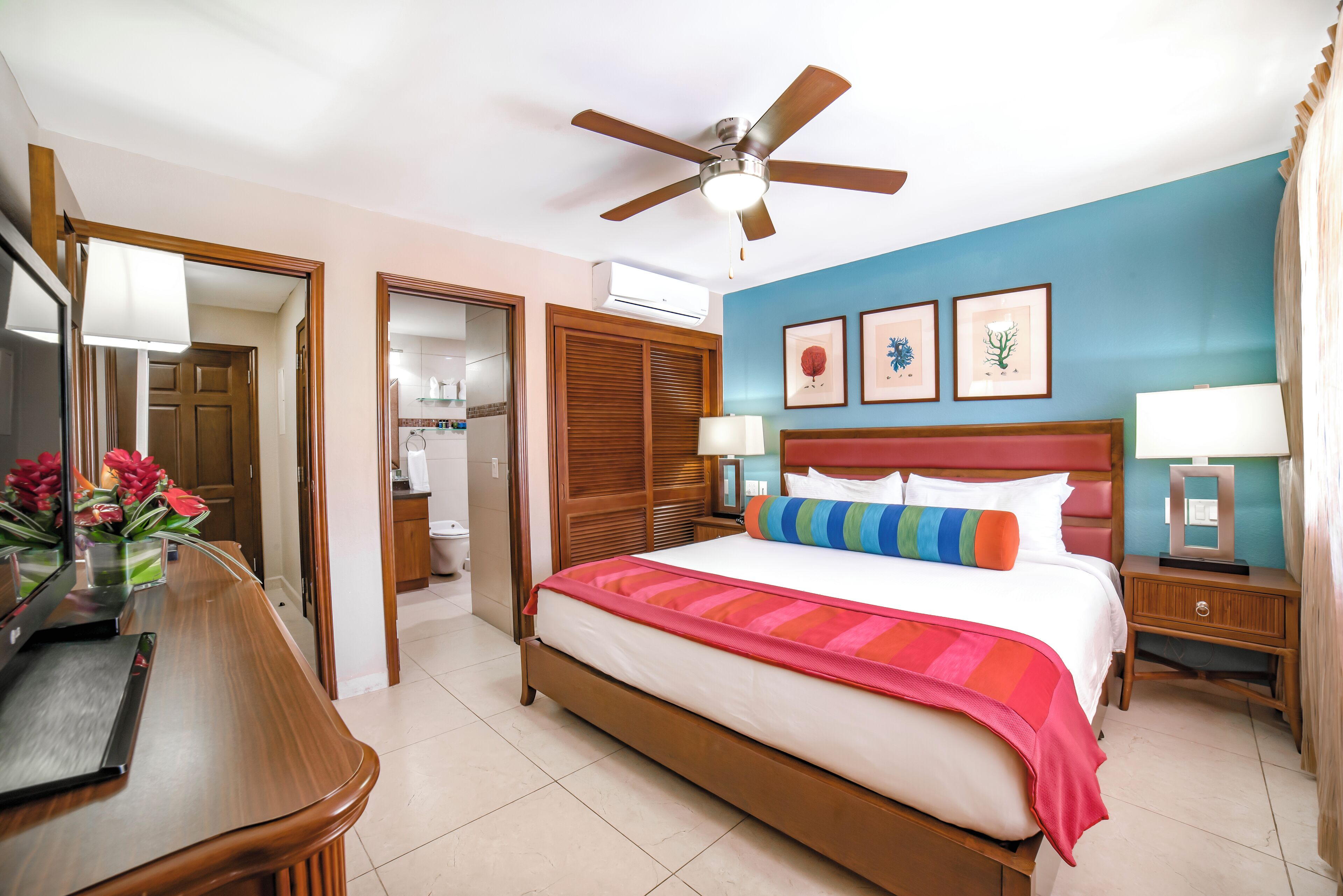 Habitación de hotel con cama, mesitas de noche, escritorio, ventilador de techo y baño visible a través de una puerta abierta.