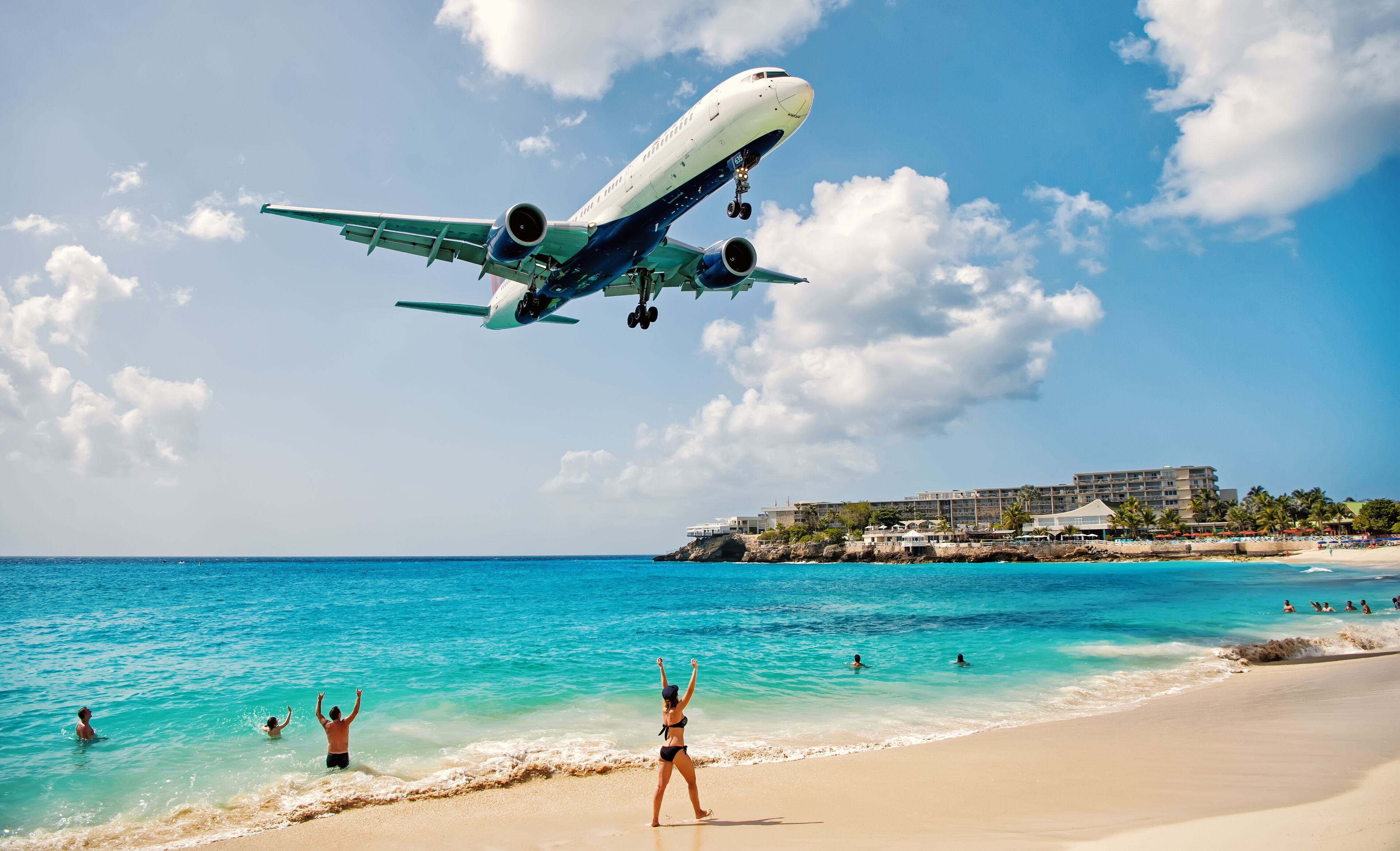 A beach with a large airplane flying overhead, clear blue water, and a resort in the background.