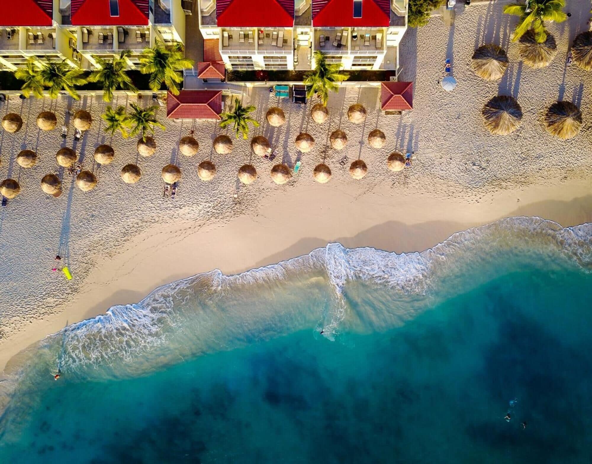 Et luftfoto af en strand med et hotel, palmer og et klart, blåt hav.