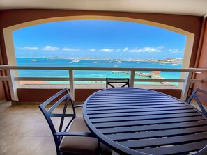 A balcony with a table and chairs, overlooking a coastal view with boats and a distant shoreline.