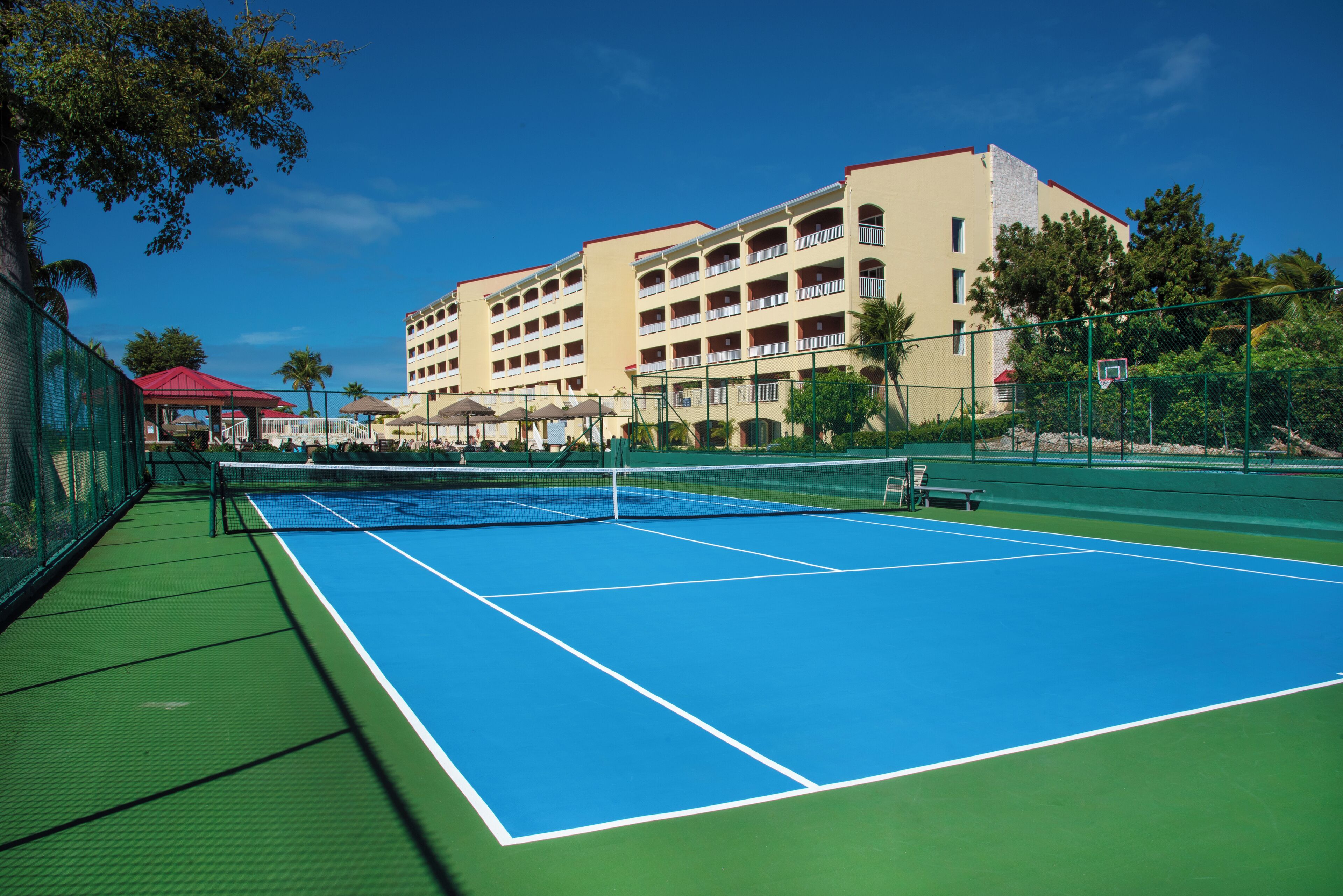 An outdoor tennis court adjacent to a hotel with multiple balconies and a red-roofed building.