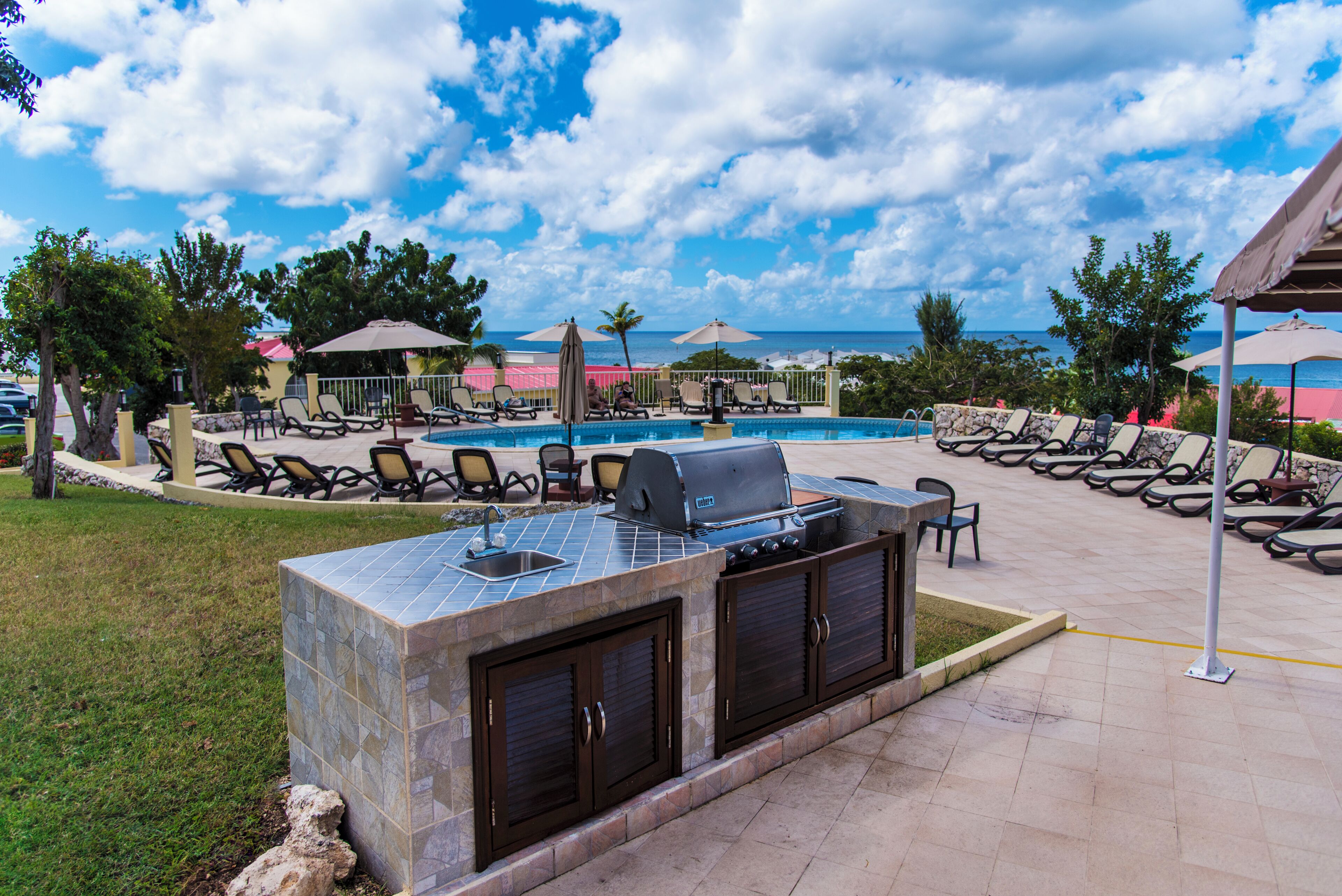 An outdoor pool area with a barbecue station, lounge chairs, and a view of the ocean.