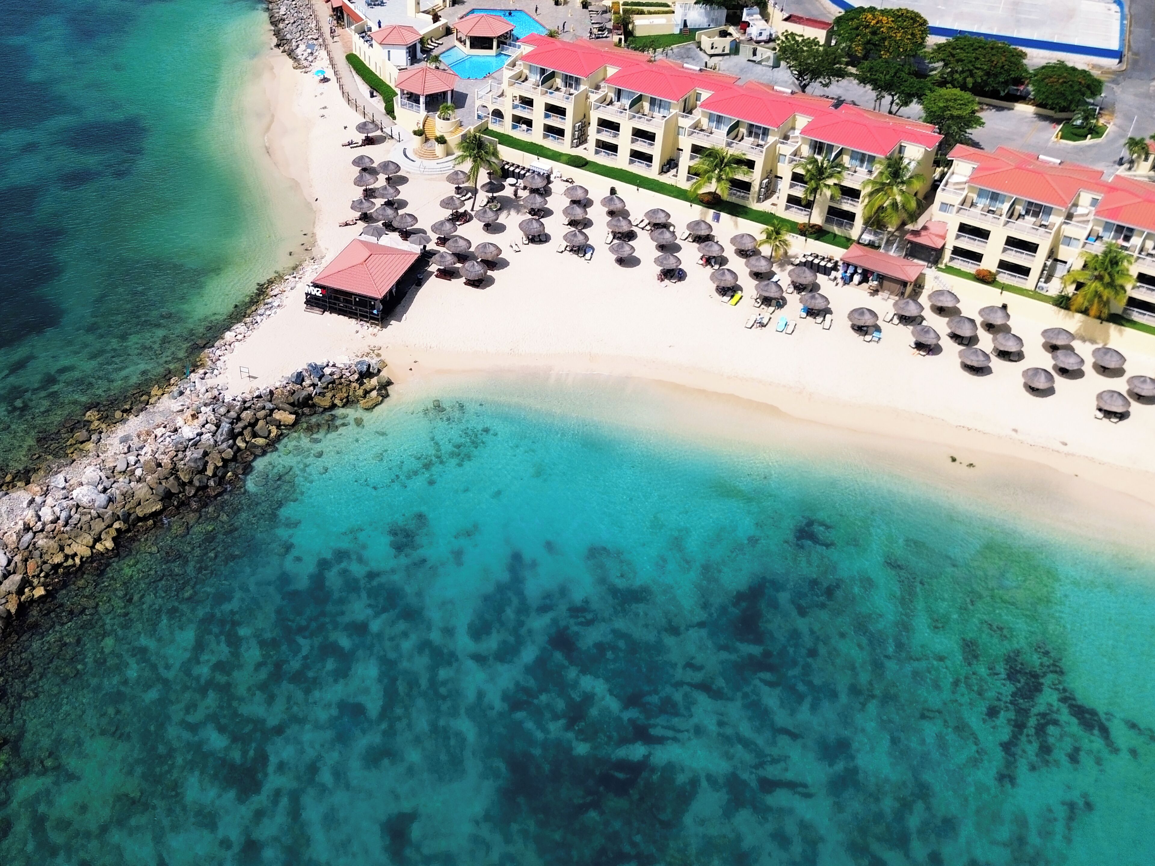 Vista aérea de un complejo turístico en la playa con un mar turquesa cristalino, un rompeolas rocoso y una hilera de villas frente a la playa con techos rojos y sombrillas de paja.