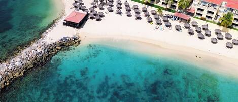 Vista aérea de un complejo turístico en la playa con un mar turquesa cristalino, un rompeolas rocoso y una hilera de villas frente a la playa con techos rojos y sombrillas de paja.