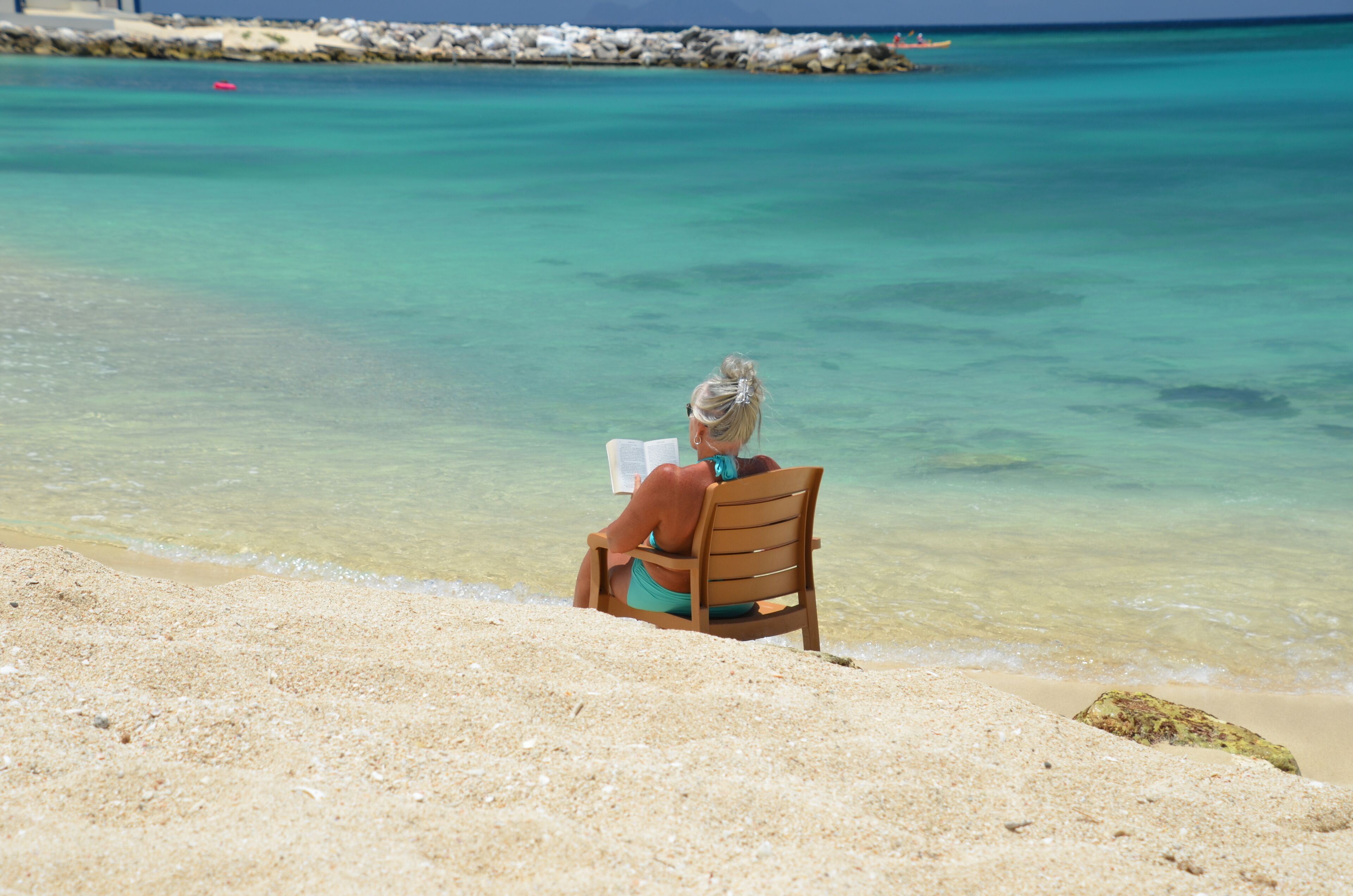 A person sitting on a wooden chair at a beach with clear turquoise water.