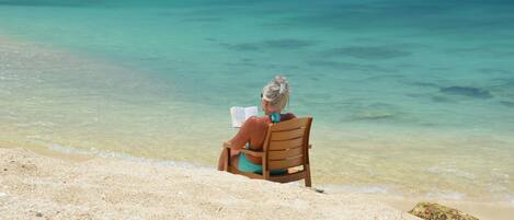 A person sitting on a wooden chair at a beach with clear turquoise water.