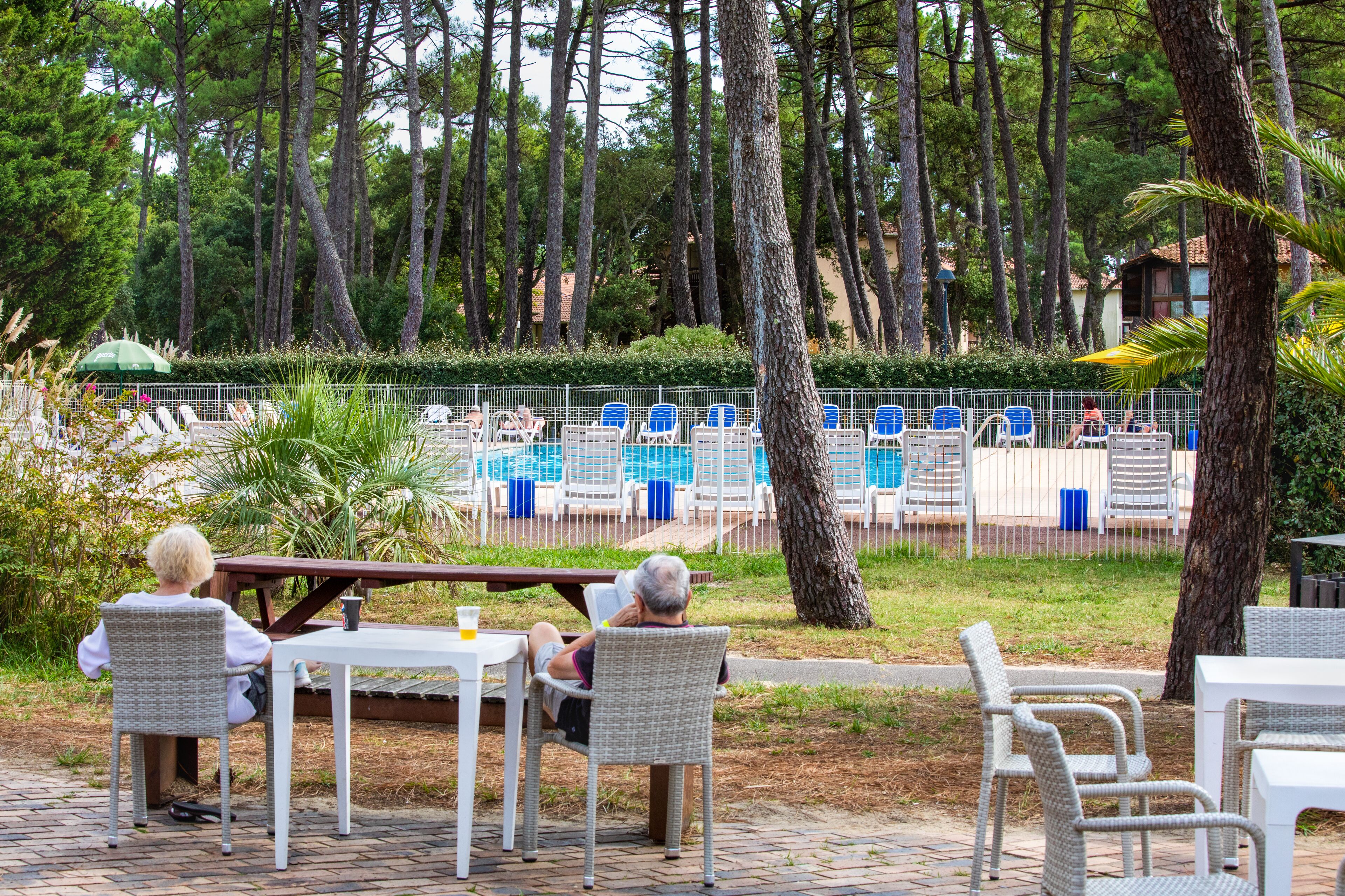 An outdoor seating area with wicker chairs and tables, a pool with blue chairs, and a fence separating the seating area from the pool.