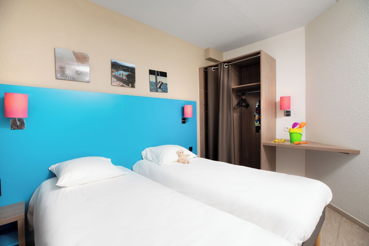 A hotel room with two beds, a blue accent wall, and a wooden wardrobe.