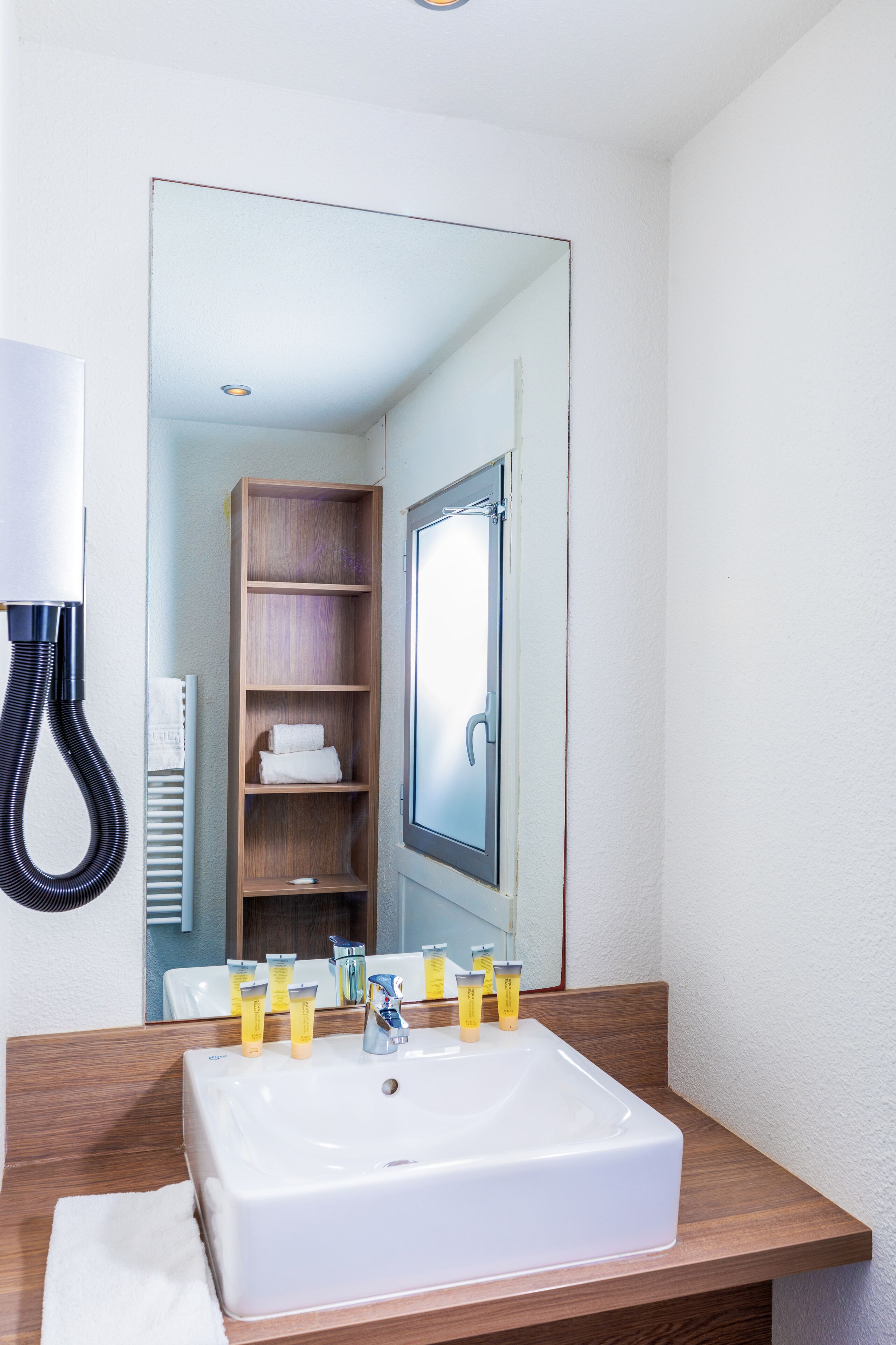 Een badkamer met een wit wastafel, houten opbergmeubel en een spiegel die een handdoekrek en planken weerspiegelt.