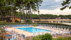 A swimming pool with lounge chairs, surrounded by tall trees and a body of water in the background. - SOWELL Family Pignada Plage (Soustons)