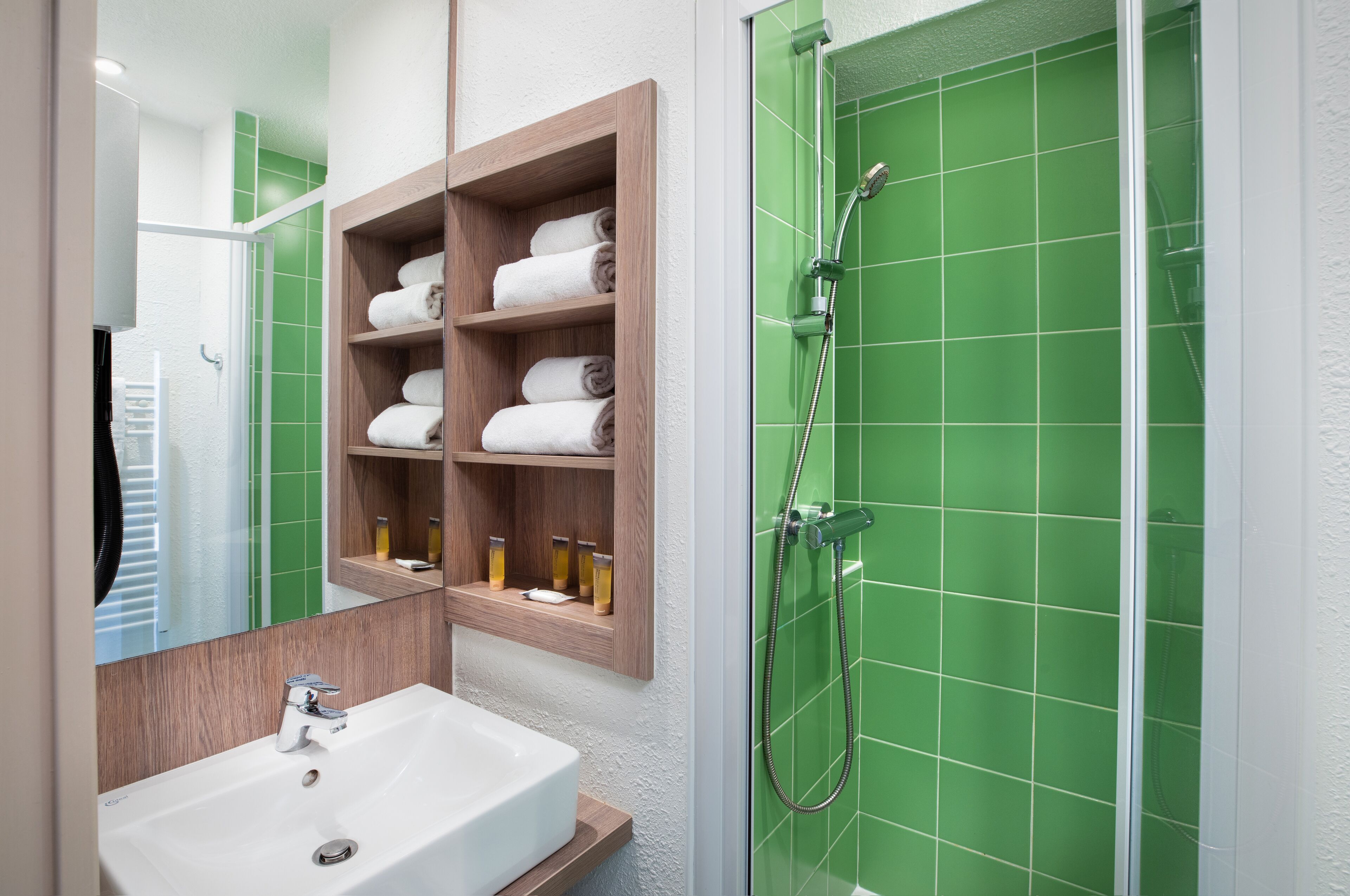 Une salle de bain avec un lavabo blanc, des étagères en bois pour les serviettes et une zone de douche carrelée de carreaux verts.