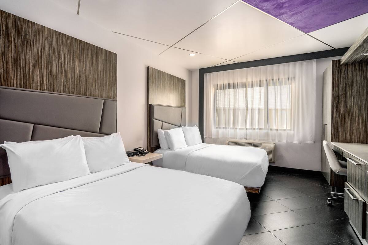 A hotel room with two beds, a desk, and a window with sheer curtains.