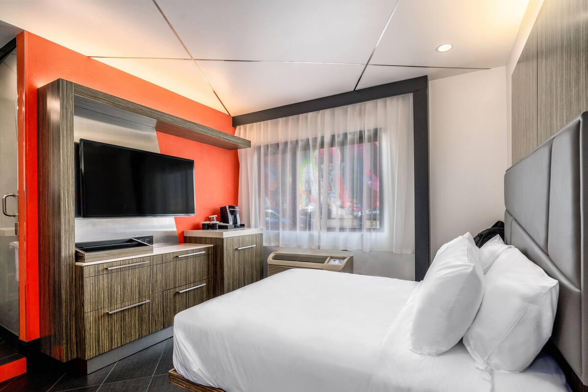 A modern hotel room with a bed, a TV, a coffee machine, and a window with sheer curtains.