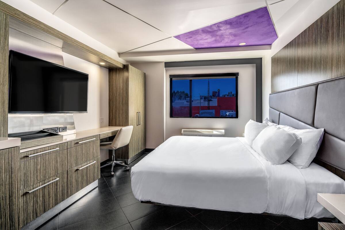 A modern hotel room with a large bed, a desk with a chair, a flat-screen TV, and a window with a city view.