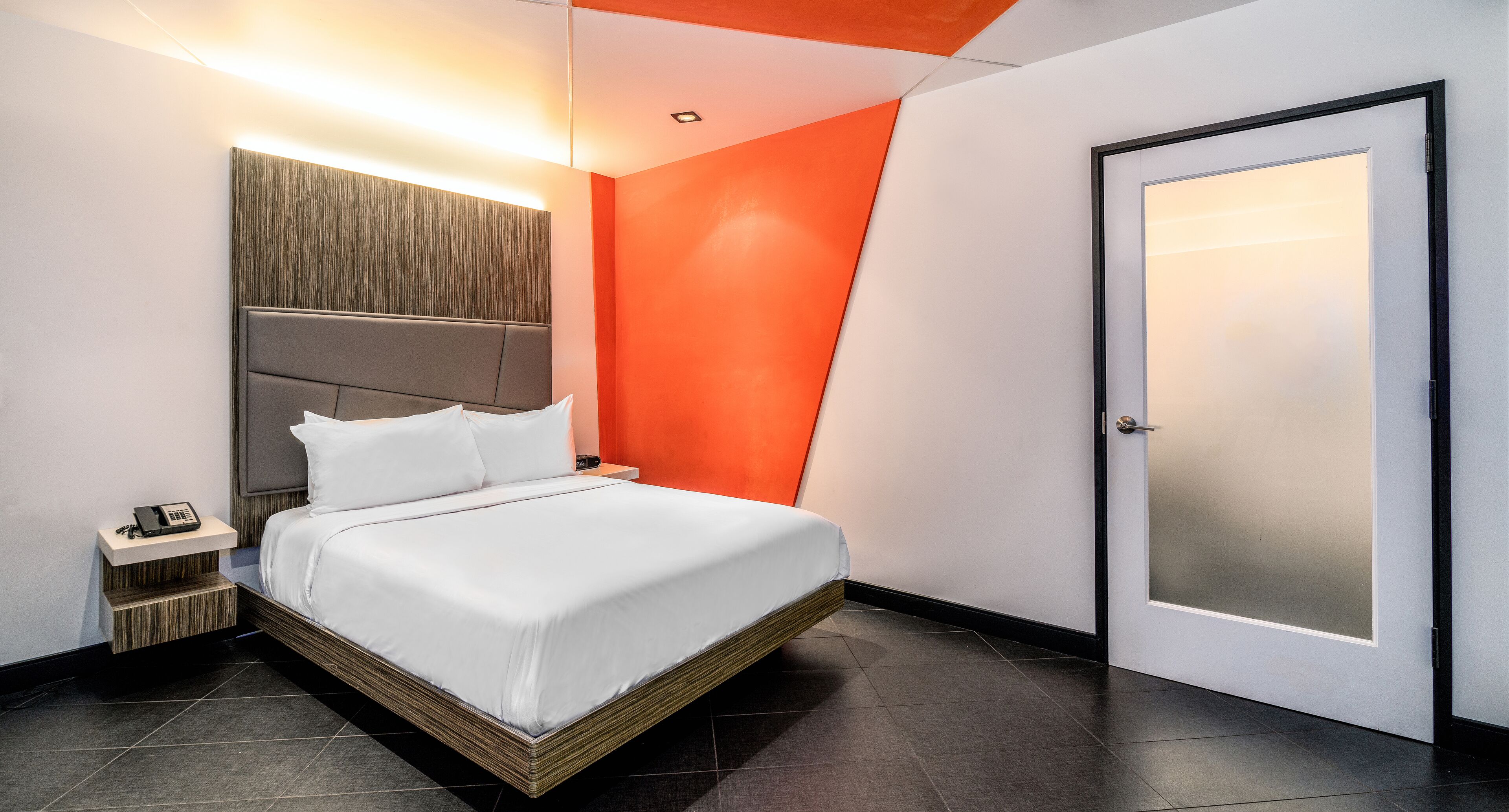 A modern hotel room with a bed, bedside table with a phone, and a door with frosted glass.