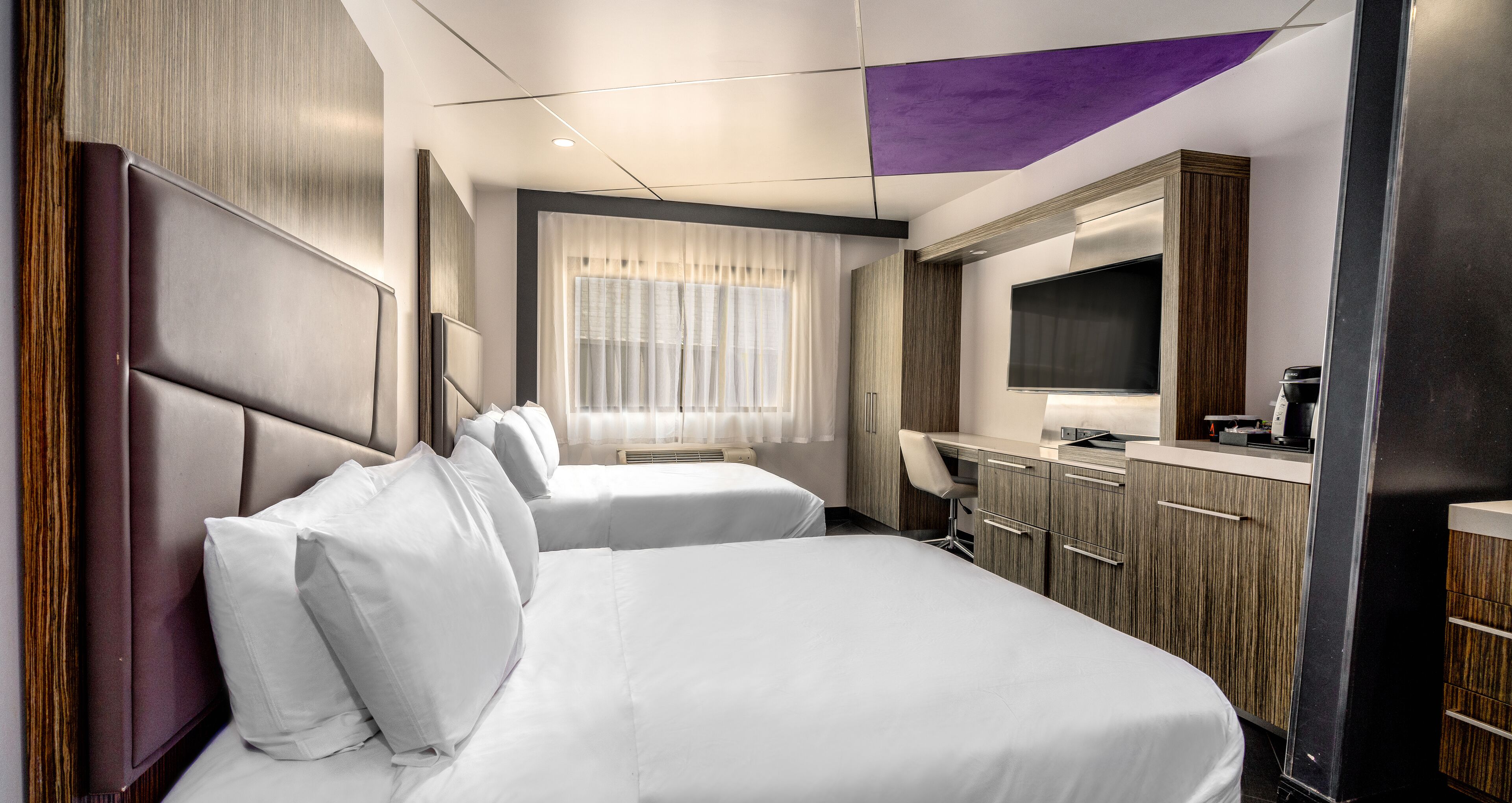 Une chambre d’hôtel avec deux lits, un bureau, un téléviseur à écran plat et une fenêtre avec des rideaux légers.