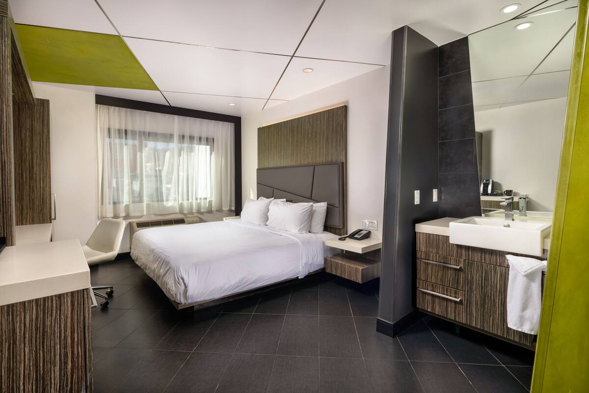 A modern hotel room with a large bed, a bedside table with a phone, a vanity with a mirror, and a window with sheer curtains.