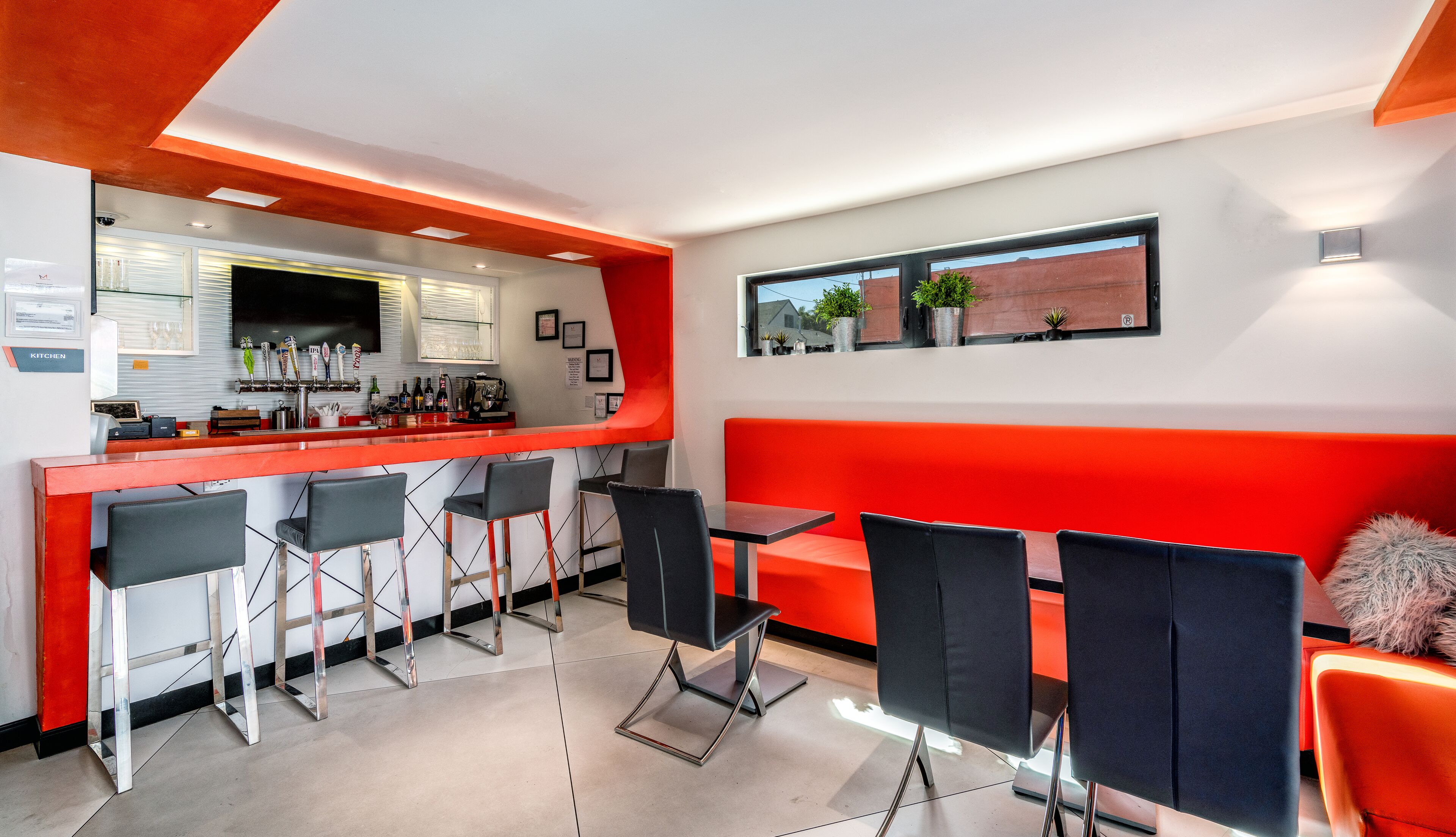 Een moderne bar met een rode toonbank, zwarte barkrukken en een zithoek met oranje en zwarte stoelen.