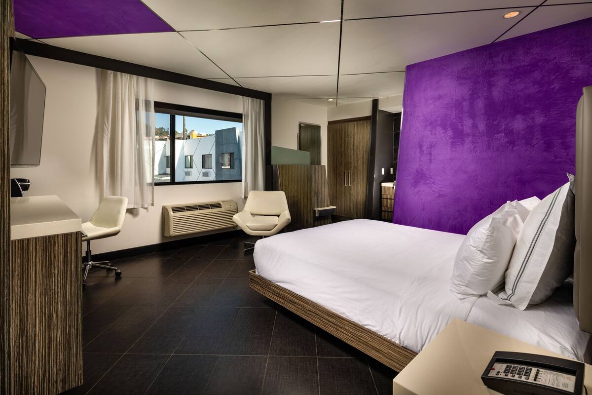 A modern hotel room with a large bed, a desk, a chair, a window with curtains, and a purple accent wall.