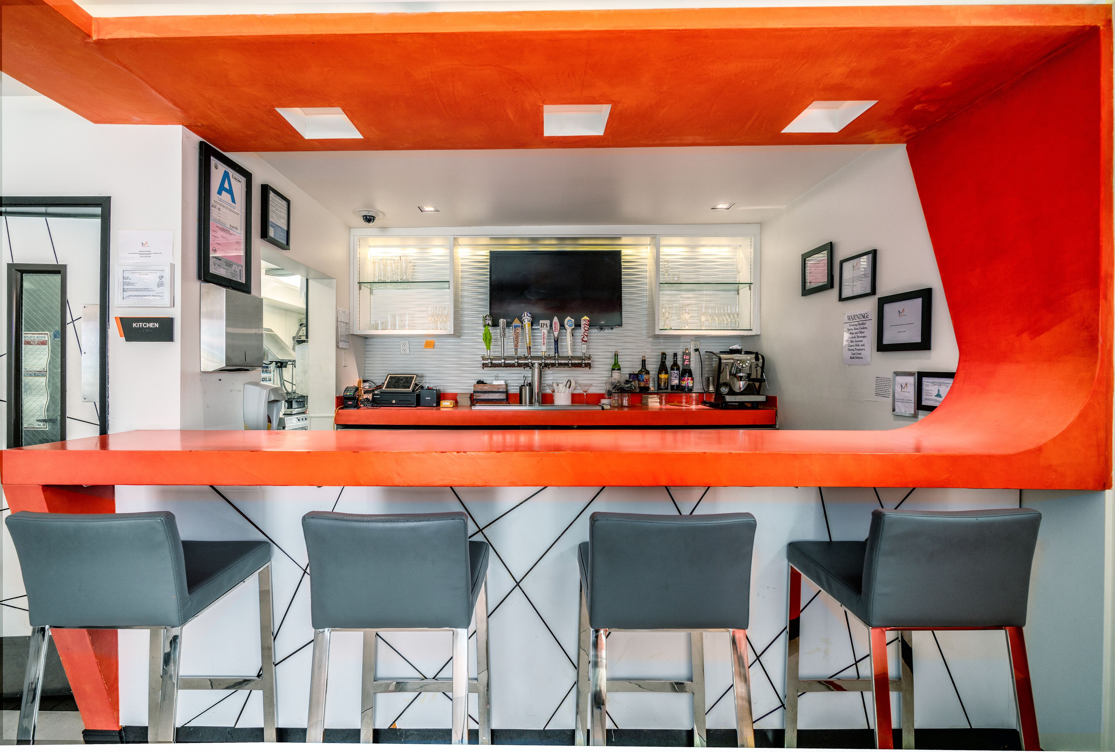 A modern bar area with a red counter, four bar stools, and a kitchen in the background.