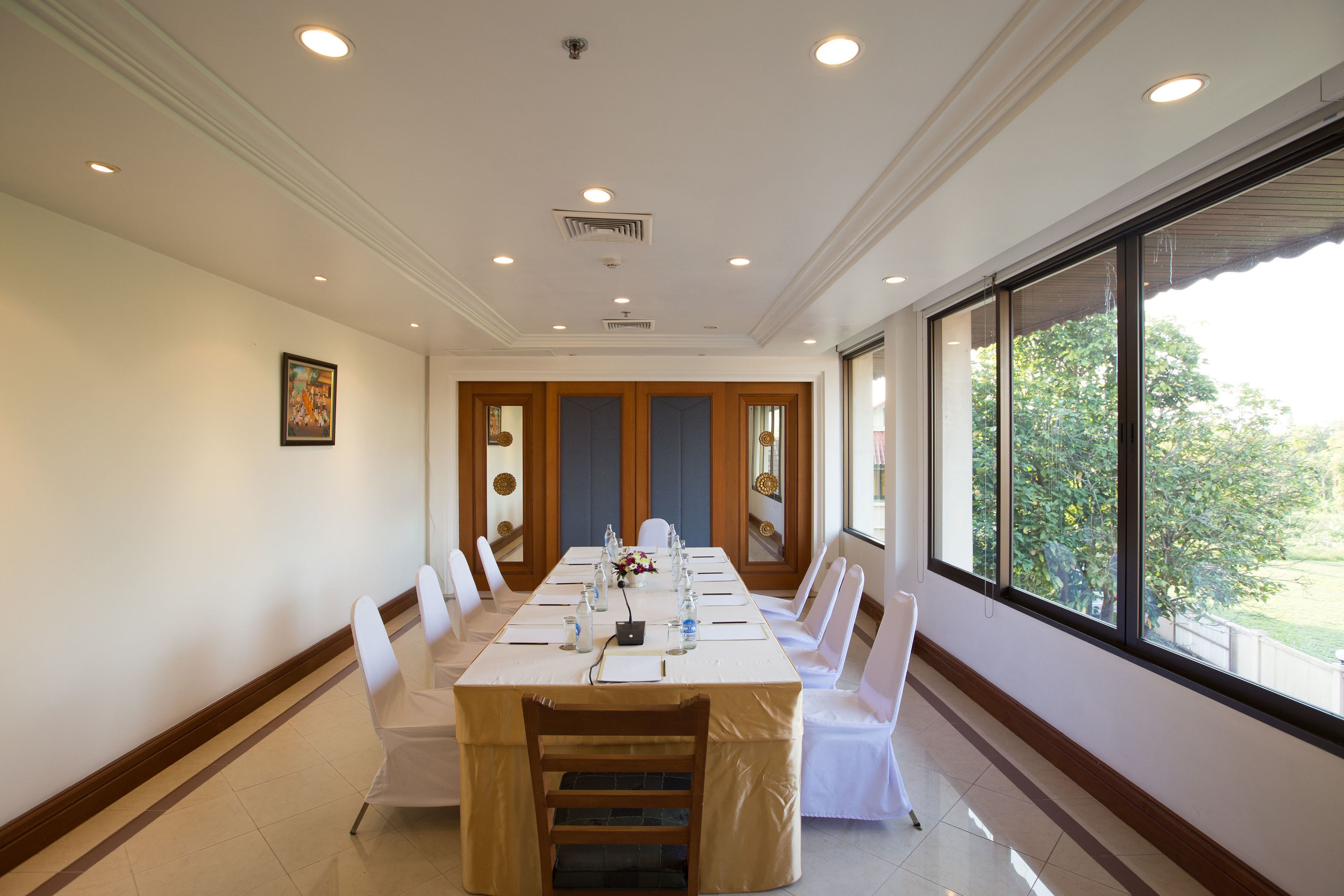 Uma sala de conferência com uma mesa comprida, cadeiras brancas e janelas amplas.