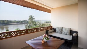 A balcony with a wooden table, a sofa with cushions, and a view of a river and greenery.