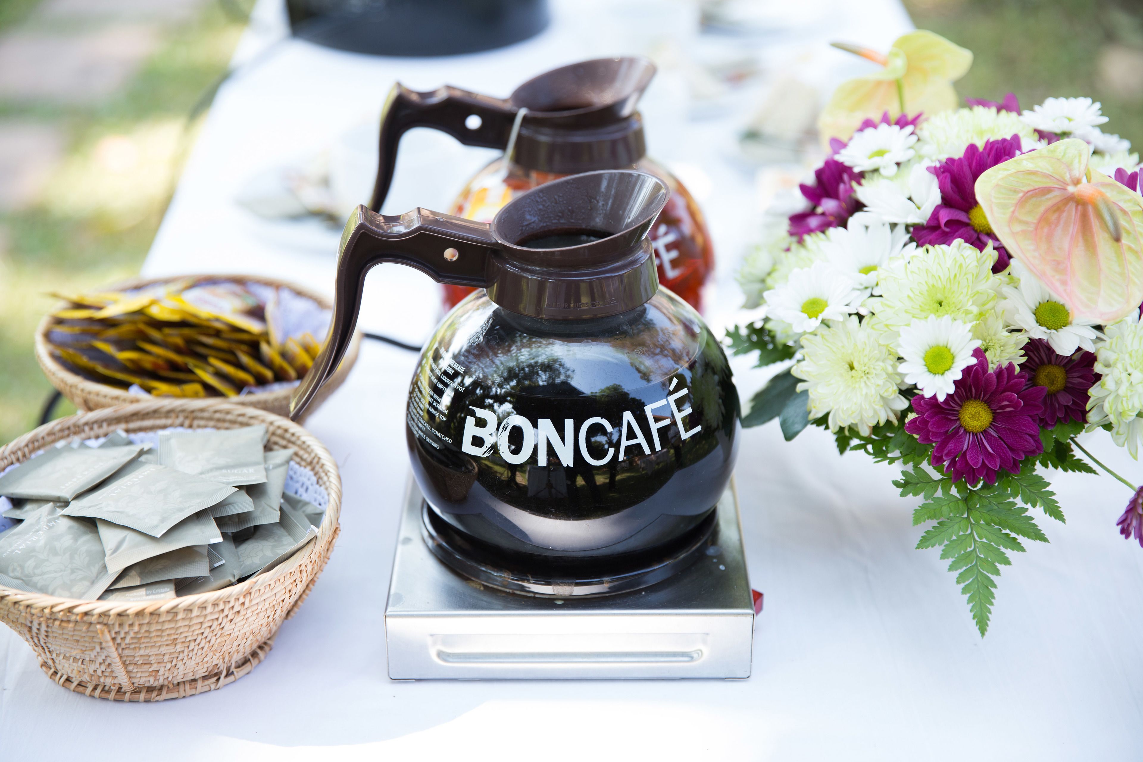 Máquina de café com a marca 'BONCAFÉ' sobre uma mesa, com um cesto de saquinhos de chá e um buquê de flores.