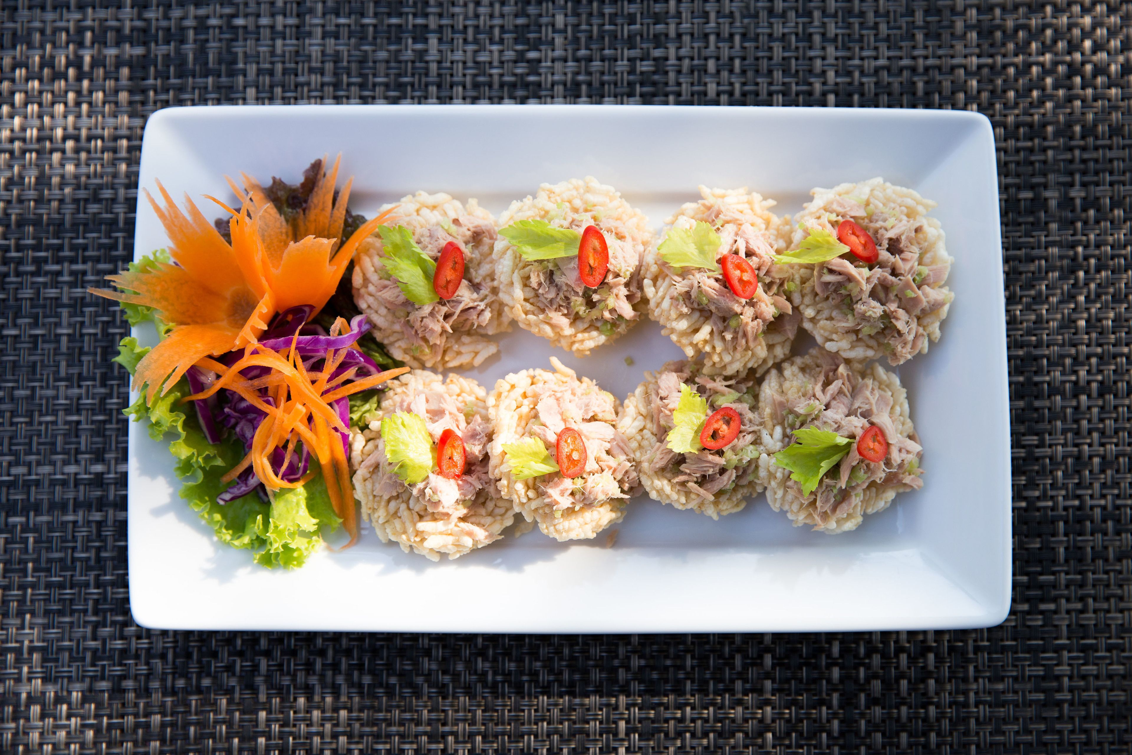 Une assiette rectangulaire blanche, sur laquelle sont disposés six amuse-gueule sphériques à base de riz, garnis de laitue, de tomates et de juliennes de carottes, accompagnés d’une salade décorative servie à part.