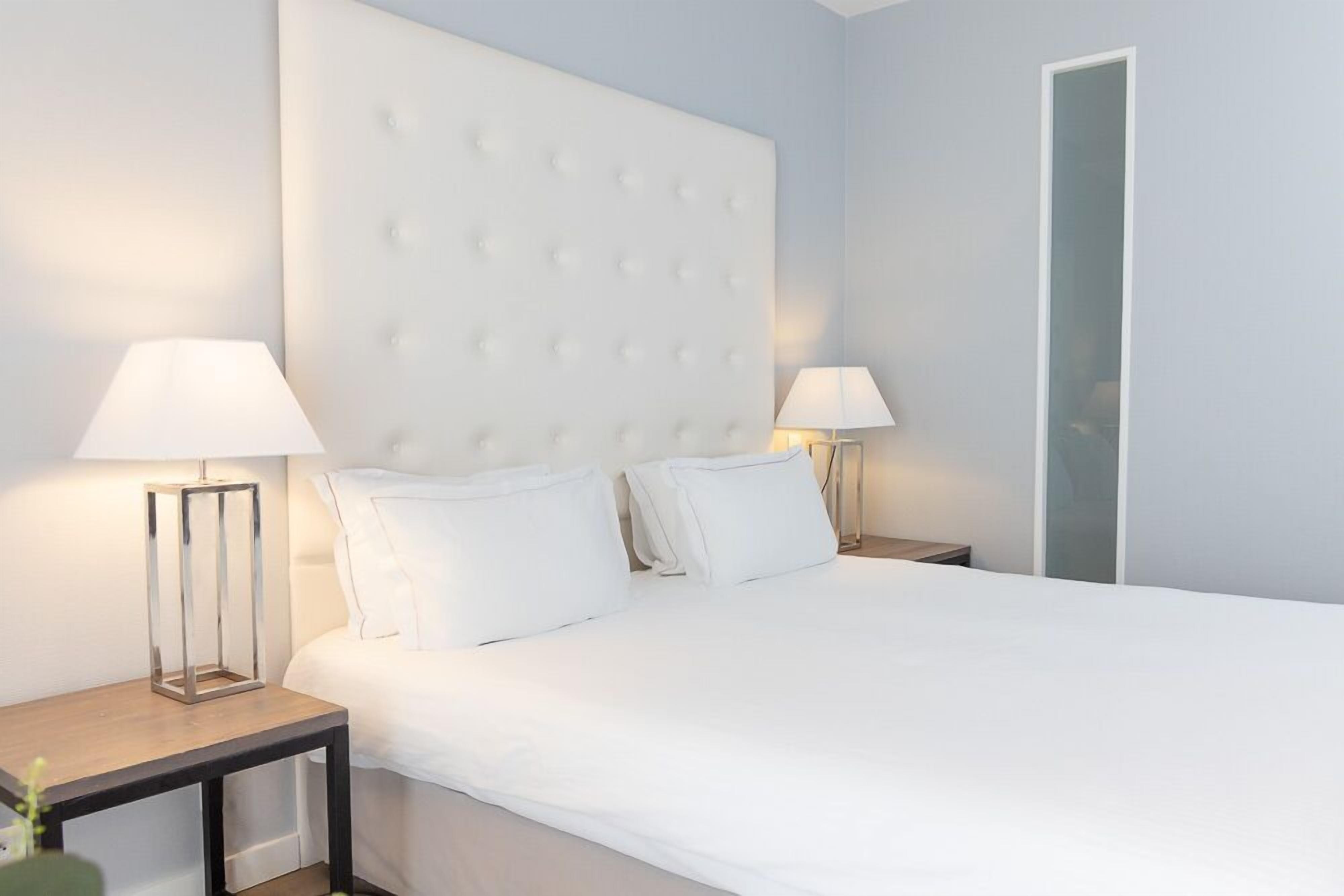 Een netjes opgemaakt bed met een gestoffeerd hoofdbord, twee nachtkastjes met lampen, en een spiegel aan de muur.